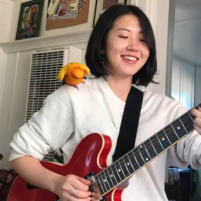 Erika Toda playing electric guitar with a colorful bird on her shoulder